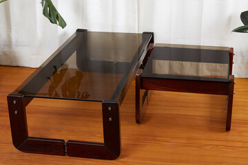 Set of vintage smoked glass coffee and side tables. Sculpted Brazilian rosewood mid-century modern furniture. Product photograph with luxurious white curtains and lively houseplants. 