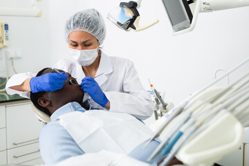 Asian woman orthodontist using tools and doing oral care inspection on african-american man patient to cure toothache.