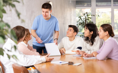 Cheerful young student discussing learning assignment with friendly coursemates gathered in classroom