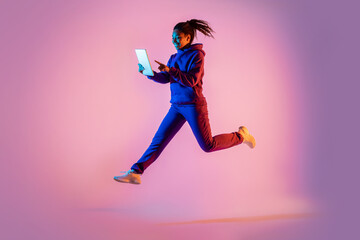 Fototapeta na wymiar Full length of young black woman with tablet computer jumping in pink neon light background, copy space