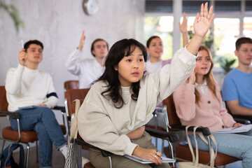 Portrait of focused diligent young Asian female student raising hand to answer while sitting in...