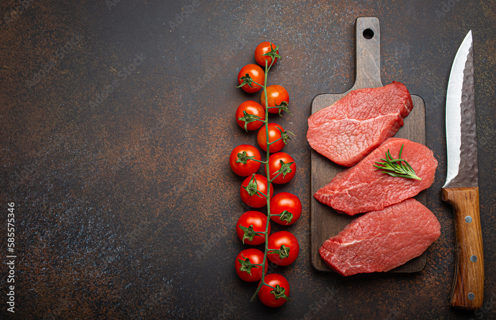 Wall mural Three raw uncooked meat beef top sirloin steaks on wooden cutting board with rosemary on wooden cutting board with cherry tomatoes branch, knife on dark rustic background top view. Copy space