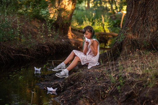 Portrait of girl with green hair sits on the river bank, holding white origami paper boat in hands
