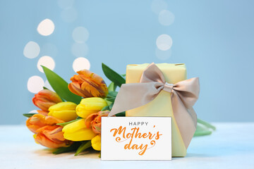 Card with text HAPPY MOTHER'S DAY, gift and bouquet of beautiful tulip flowers on table against...