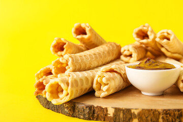 Board with delicious wafer rolls and boiled condensed milk on yellow background