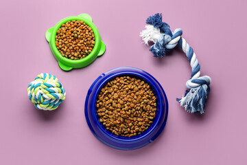 Bowls with dry pet food and toys on color background