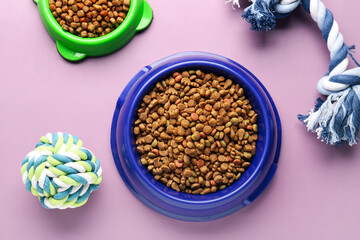 Bowls with dry pet food and toys on color background
