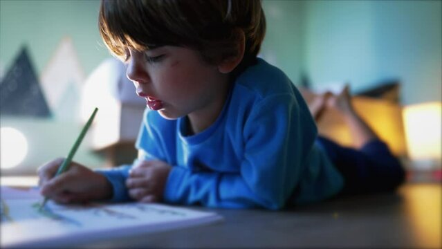 One small boy laying on floor coloring on paper. Child doing school activity wearing pajamas in morning. Kid studying homework in bedroom