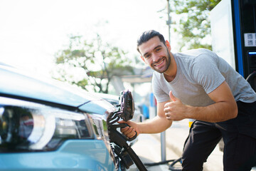 Naklejka premium Handsome Asian young man holding an EV plug connector and attaching onto the car, man plugged in the EV charging connector to the vehicle.