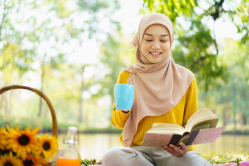 Happy cheerful Asian beautiful muslim woman sitting on the ground at the park and drinking a cup of...