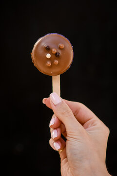 Hand Of Woman Gently Holding Delicious Macaron In Chocolate Icing On A Stick On Black Background.