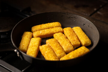 Breaded fish sticks in a frying pan. Preparation of frozen fish sticks. Fast food. Dark background.