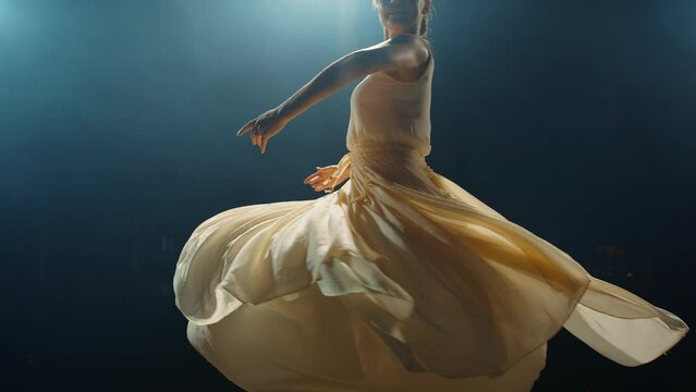 Graceful ballerina dances in light flowing dress on stage in pointlight in stage smoke. Ballet dancer performs dance elements of classical ballet