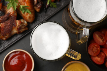 Mugs with beer, delicious baked chicken wings and sauces on dark grey table, flat lay
