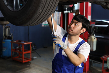 Man technician car mechanic in uniform check or maintenance a lifted car service at repair garage station. Worker holding wrench and fixing vehicle with flashlight. Car center repair service concept