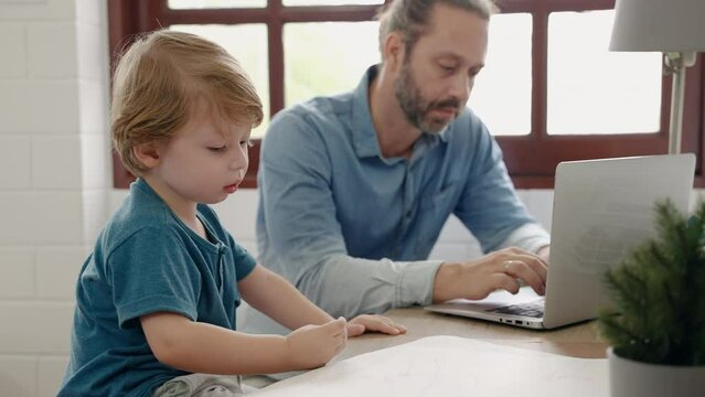 Father sitting at work using tablet while son drawing and painting, family relations concept
