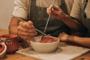 A young couple, a man and a woman, are painting a bowl with paints in a pottery workshop. Close-up hands.