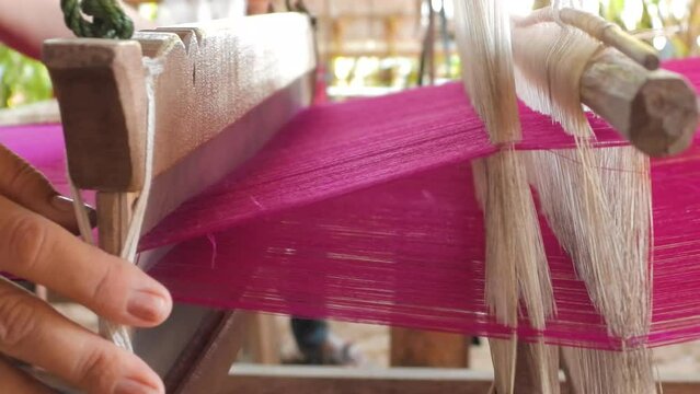 Close up of a female hand on a loom to produce Fuchsia Pink Thai silk, by alternating silk threads bobbin for weaving left and right, Traditional silk production on the Thai countryside.