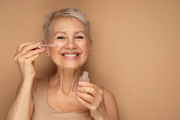 Happy senior lady applies cosmetic oil serum on face takes care of skin and smiles broadly enjoys beauty treatments