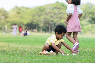 Fototapeta premium Little African boy and girl wearing bunny ears pick up Easter egg in garden, Easter eggs hunting outdoors