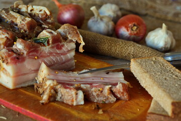 Pieces of salty lard cooked with spices on wooden cutting desk, knife and slices of grey bread on blurred background of onion and garlic bulbs. Salo - Traditional food of Ukrainian and Russian cuisine