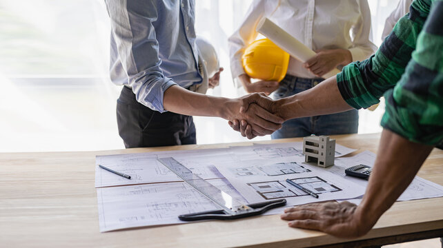 Yellow hard hat on table and house design print design with construction team handshake greeting start new project contract plan in office center at construction site partner and contractor
