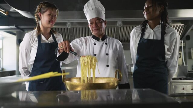 Happy Asian senior man chef in uniform standing teaching and showing spaghetti food cooking with diverse students in kitchen room. 