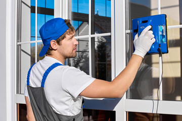 Man in overalls with white gloves presses a button to start the robot to wash the window. Inclusion of a modern square-shaped robot for washing glass