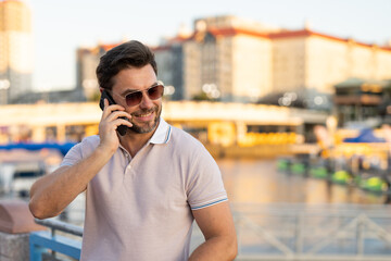 Portrait of handsome man chatting on phone outdoor. Stylish man talking on phone dressed in polo. Fashion male posing on the city street. Urban style. Business phone conversation.