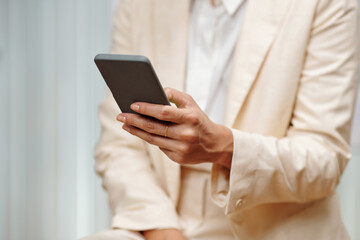 Hand of young woman in beige blazer texting in mobile phone or scrolling through contacts while standing in front of camera in office