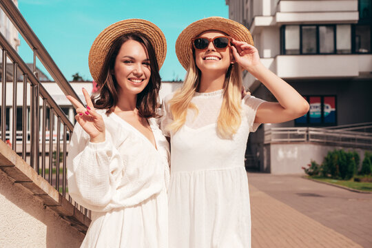 Two young beautiful smiling hipster female in trendy summer white dress and straw hats. Sexy carefree women holding and drinking fresh vegetable cocktail smoothie drink in plastic cup with straw