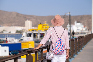 Rear view of mature woman with summer hat looking at ferry boat departure in the port - weekend vacation travel destination concept