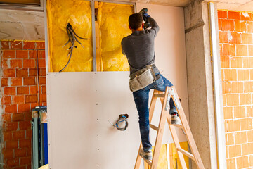 Worker on ladder uses hand drill, ACU power tool and twists screws on rock wool isolated gypsum dry...