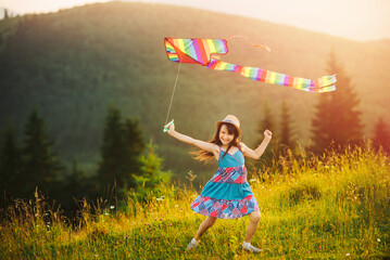 Little girl runs colored striped rainbow kite at sunset on summer day. Happy summer vacation and healthy lifestyle concept. Mountain landscape with coniferous trees.