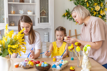 A happy mom with her adult daughters paint and decorate Easter eggs at home in the kitchen. Preparation for the Easter holiday