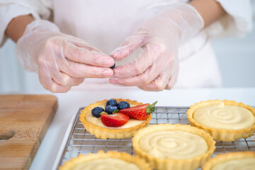 close up of hands in cooking gloves Baker adding blueberries strawberry fresh fruit to a tart on white table in Kitchen. housewife baker wear apron making fruit tart. homemade bakery at home.