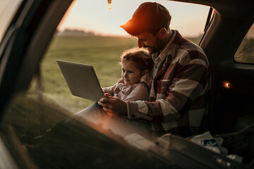 A man sitting in a car truck during the sunset with his daughter sitting on his knees, both using a...