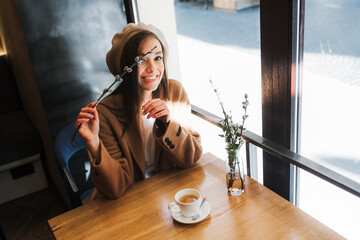 Young attractive woman sitting in tea room at table by the window. Twig with willow catkins in hand.