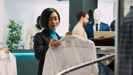 Asian customer looking at formal wear shirt in retail shop, using fashionable brands to increase wardrobe. Female adult buying fashion boutique merchandise, small business concept.