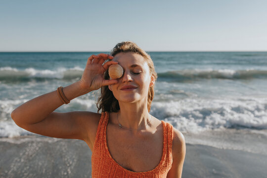 Smiling woman holding seashell over eye in front of sea