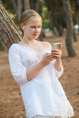 Teenage girl in white clothes and headphones holding mobile phone and standing near tree on the background of forest