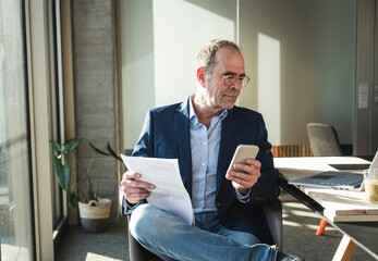 Mature businessman using smart phone sitting with document in office