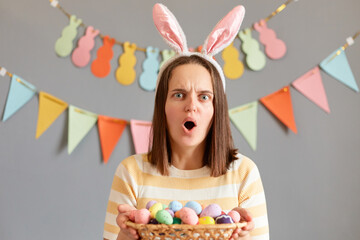 Portrait of woman wearing striped shirt celebrating easter isolated over gray background, holding basket with eggs, looking at camera with surprised expression.