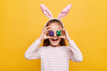 Portrait of funny little girl with rabbit ears with covering her eyes colored purple and green eggs having fun smiling happily posing isolated on yellow background