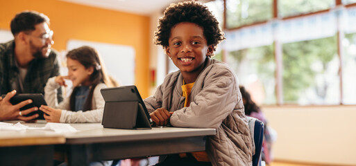 Boy smiling at the camera while sitting with a digital tablet in a classroom
