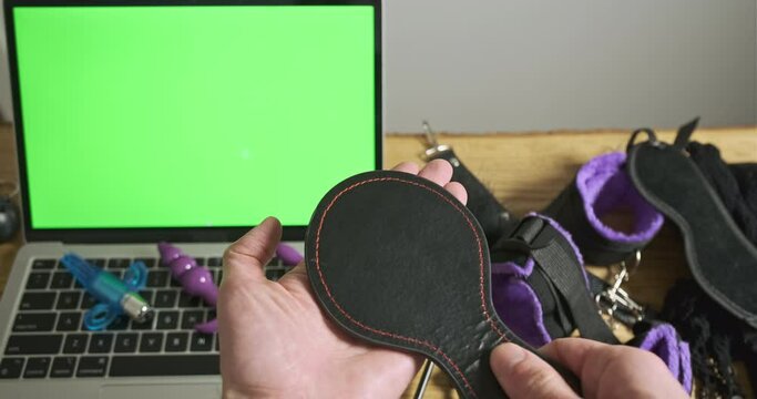 Man's hands check the bdsm spatula for spanking. Laptop with green screen, sex toys lie on the table. Top view, close-up.