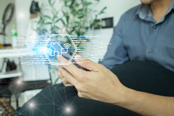 A man is sitting and watching the operation status of home devices through an application in his mobile phone with a hud holographic graphic on the front, the concept of future technology Smart Home.