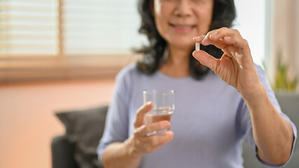 A retired Asian woman holding a glass of water and a pill, taking a medicine in her living room.
