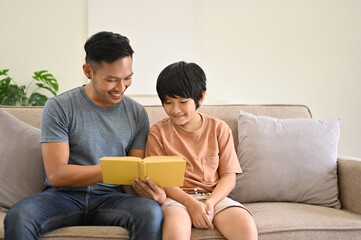 Fototapeta premium Happy and kind Asian dad is telling a story to his son on a sofa in the living room