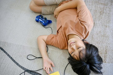A happy cute Asian little boy laying on the living room floor with video game joysticks.